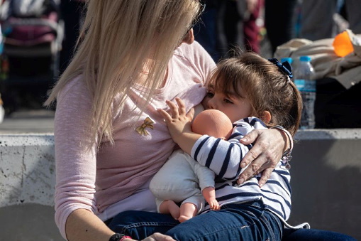 Zweijähriges Mädchen mit Puppe auf dem Arm trinkt an der Brust ihrer Mutter in einem öffentlichen Park