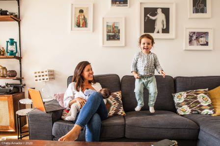 Stillende Mutter sitzt auf dem Sofa mit ihrem Baby während ihr älteres Kind auf dem Sofa herumhüpft