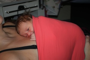 Baby liegt Haut an Haut auf dem Bauch der Mutter, sie beide haben zusammen ein rotes Tuch an.